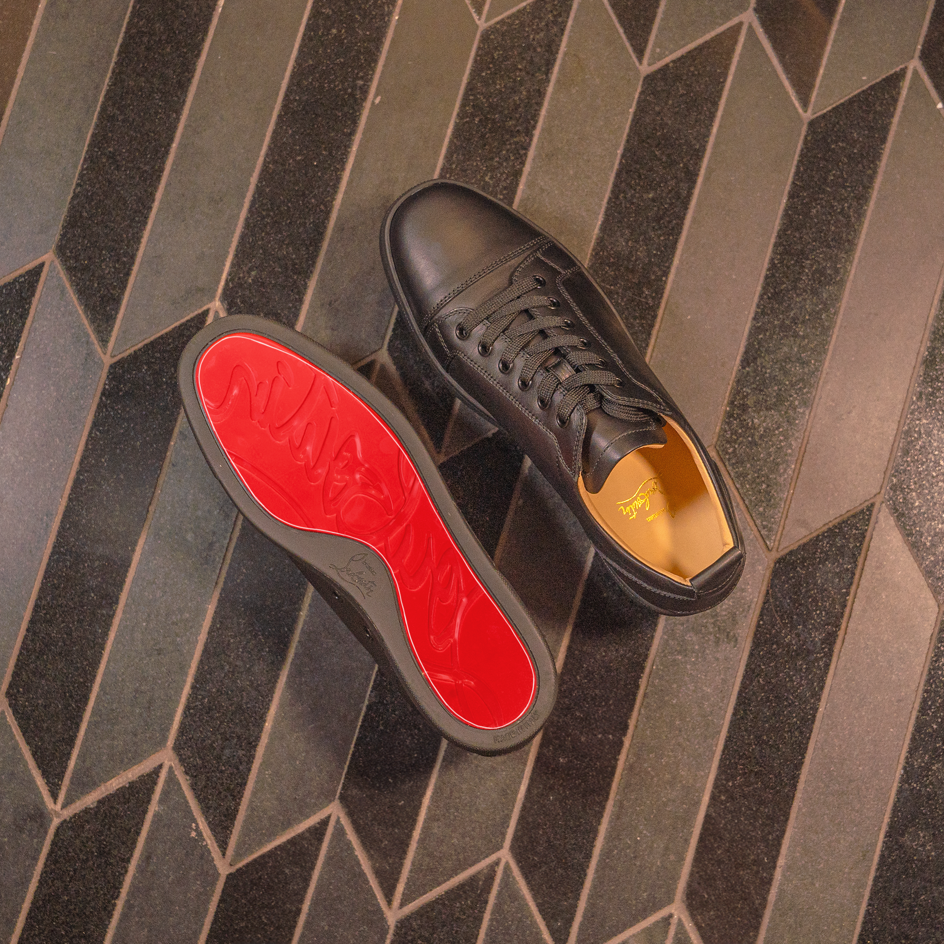 Christian Louboutin Sneaker Sole Guard with red soles on a dark, herringbone-patterned floor.