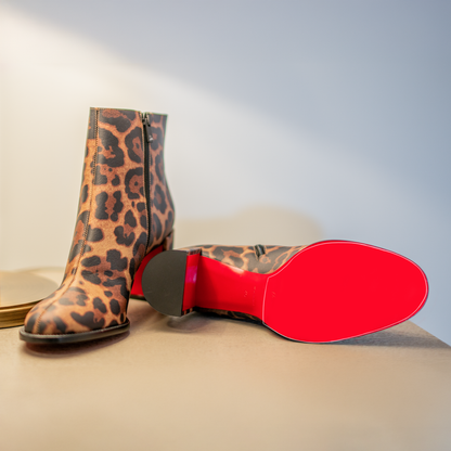 Leopard print ankle boots with iconic red soles: one stands upright, the other lies on its side, on a light surface. They feature a Sole Guard for elegant and stylish Christian Louboutin footwear. Product: Christian Louboutin Block Heel Boots Sole Guard.