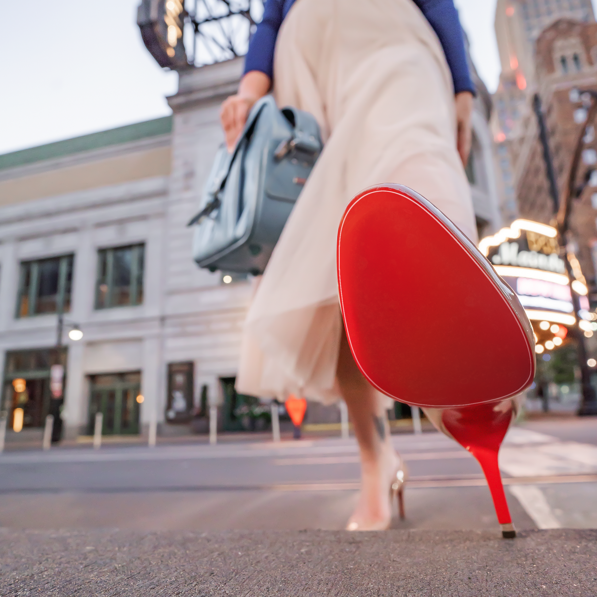 A person confidently strides in sleek attire, featuring a skirt and a blue bag, while wearing Christian Louboutin Pumps Sole Guard 3x Pack with standout red soles against an urban backdrop.