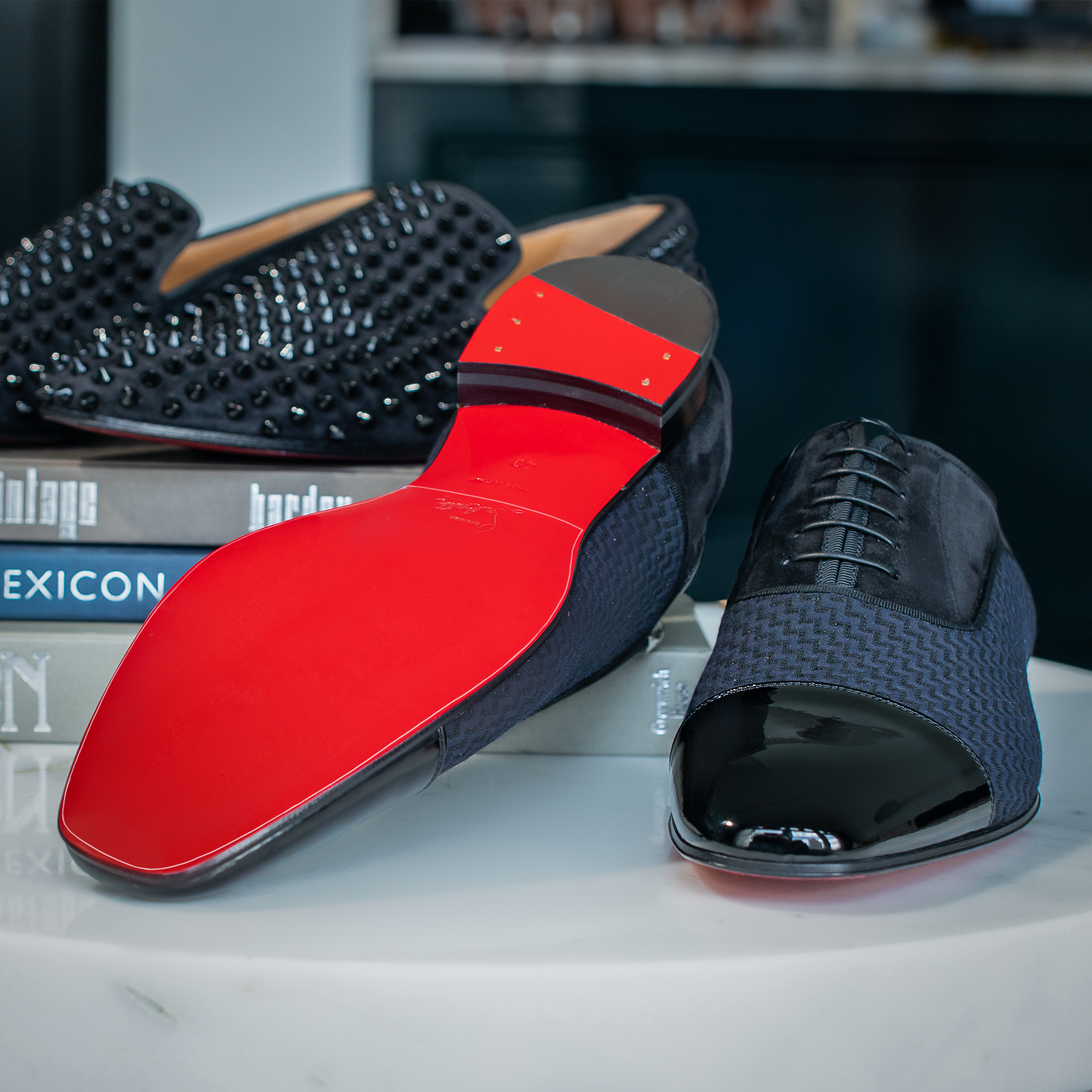 Two pairs of luxury Christian Louboutin Oxfords Sole Guard men's shoes, one with stud embellishments and iconic red soles, are elegantly displayed on stacked books.