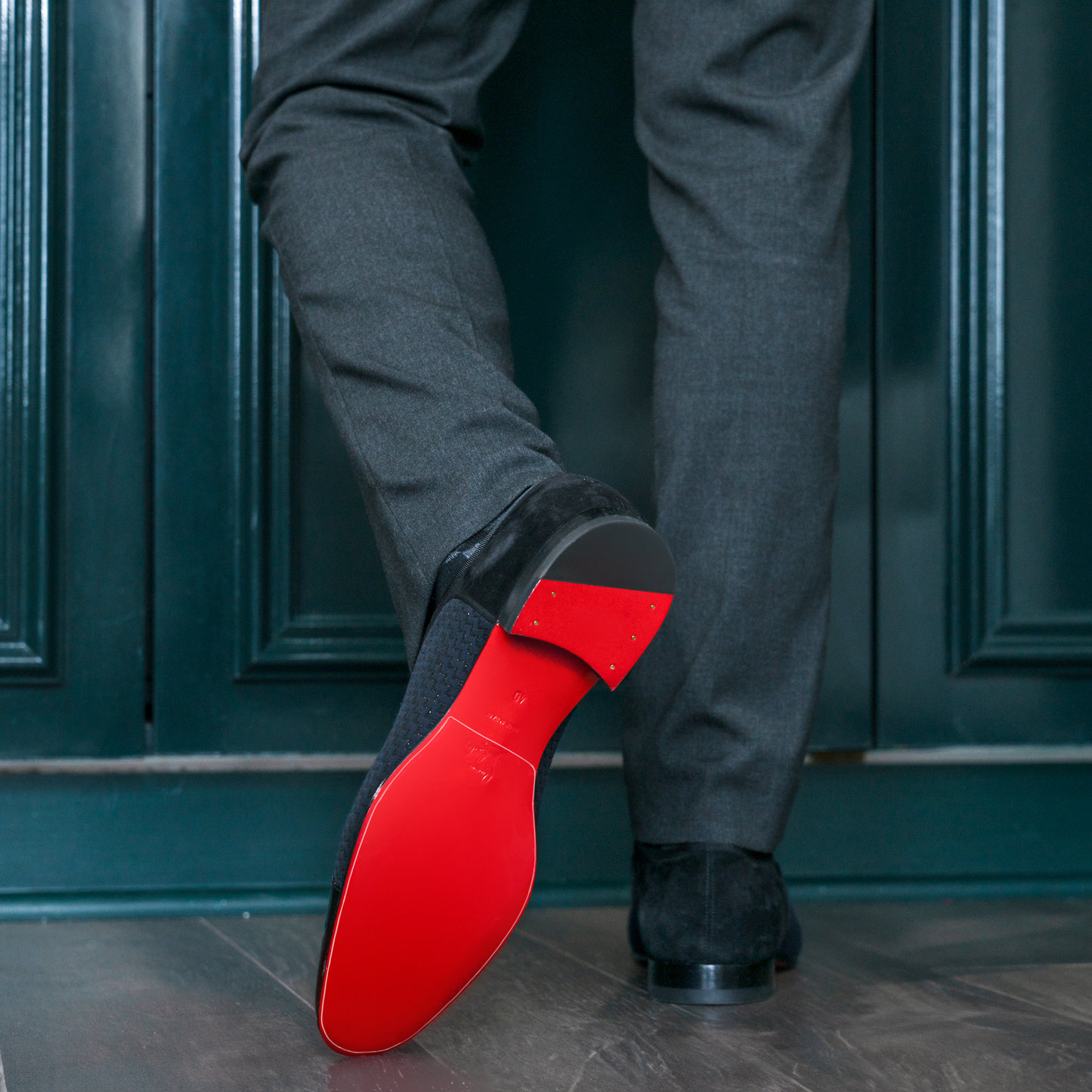 A man in gray pants strides confidently past a dark green door, his foot adorned with Christian Louboutin black Oxfords featuring their iconic red sole, protected by an invisible Sole Guard.