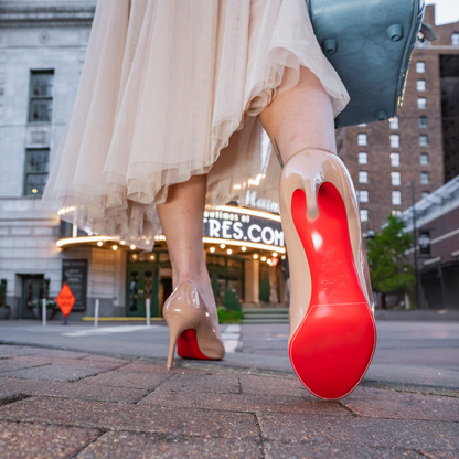 A woman in a white dress strides gracefully in Christian Louboutin pumps with red soles, each step safeguarded by the Christian Louboutin Pumps Sole Guard 3x Pack, outside a theater on a bustling city street.