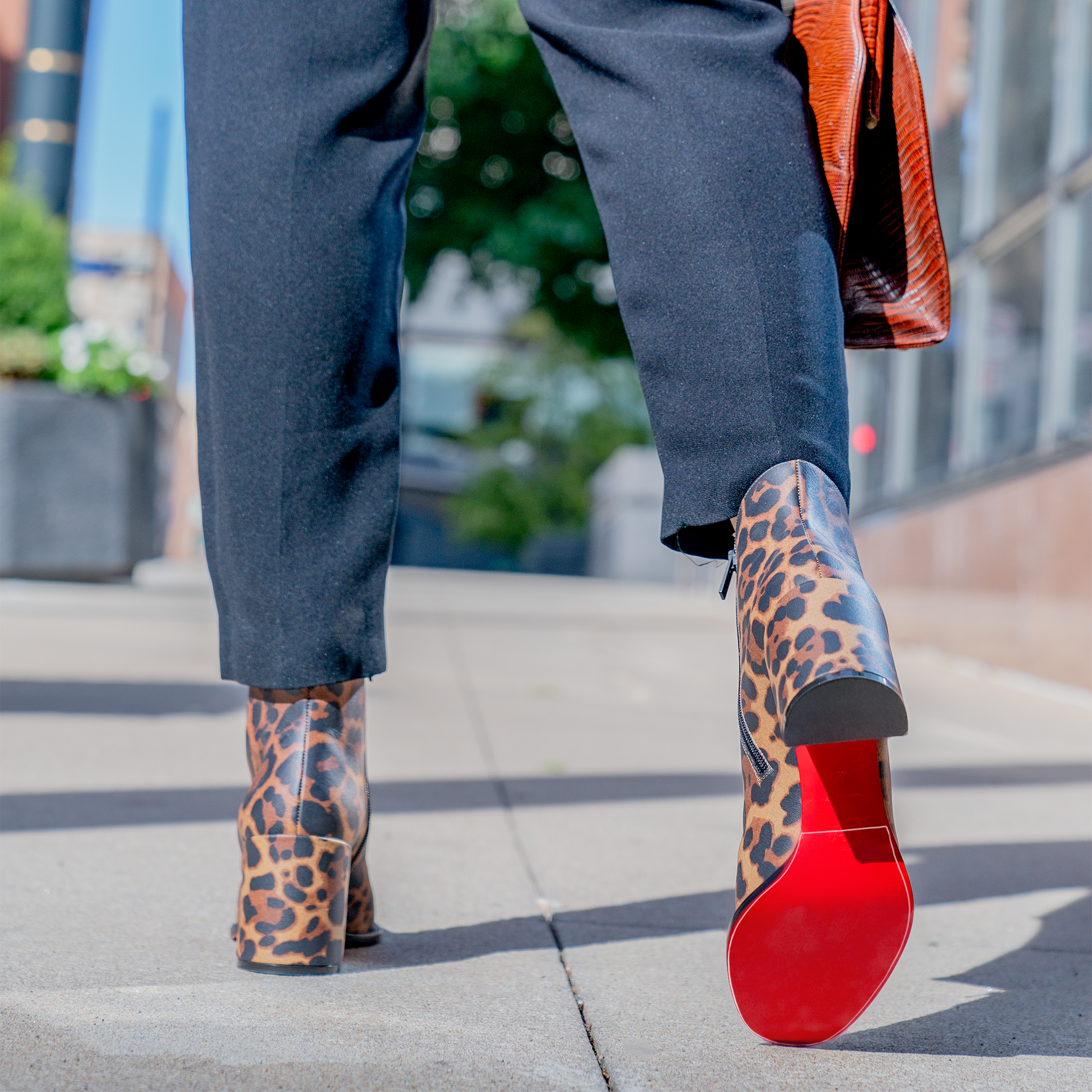 Close-up of a person walking on a sidewalk wearing Christian Louboutin Block Heel Boots with leopard print and signature red soles, complemented by black pants. The boots are well-preserved with Sole Guard protection.