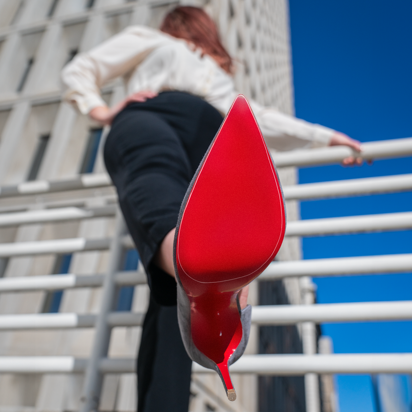 A low-angle shot showcases a woman in black pants wearing the iconic Christian Louboutin Sole Guard 3 Pack heels, with their signature red soles strikingly contrasting against a modern building under the clear blue sky.