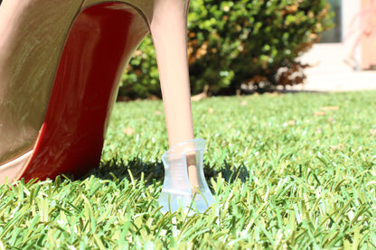 A high heel with "High Heel Protectors" gracefully stands on grass, against a backdrop of bushes.